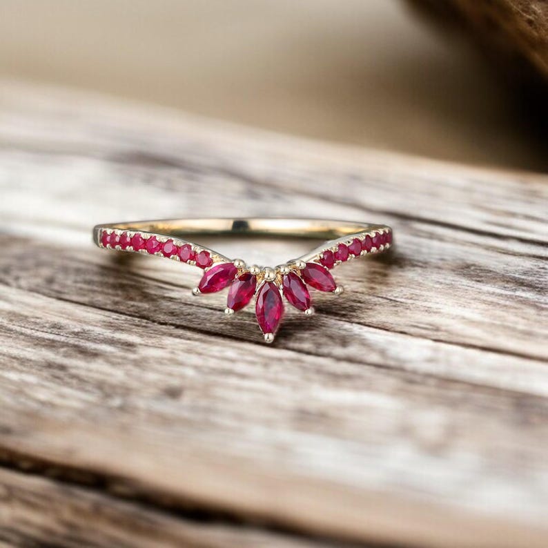 Op de afbeelding: Een goudkleurige ring met een gebogen ontwerp, bezet met kleine rode edelstenen langs de band en grotere, marquise-geslepen rode stenen die een centraal bloem- of bladachtig patroon vormen. De ring wordt gepresenteerd op een houten oppervlak.