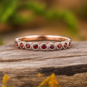 Puede incluir: Un anillo de oro rosa con una hilera de pequeñas piedras preciosas granates redondas de color rojo intenso. Cada piedra está engastada en un bisel con detalles milgrain, creando un diseño texturizado continuo en la parte frontal del anillo.