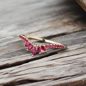 Op de afbeelding: Een goudkleurige ring met een gebogen ontwerp, met een rij kleine, ronde rode edelstenen en een cluster van grotere, marquise-geslepen rode edelstenen. De ring ligt op een rustiek houten oppervlak.