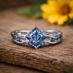 Puede incluir: Un anillo de tono plateado con una piedra preciosa azul central en forma de cometa montada en garras. La banda presenta un intrincado diseño de metal en forma de enredadera que rodea el dedo, adornado con piedras azules más pequeñas a los lados.