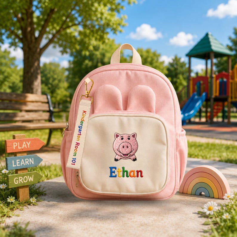 May include: A light pink backpack featuring two rounded ears on top, a cream-colored front pocket with an embroidered pig face, and the name Ethan in multicolored letters. A cream fabric tag attached to the zipper reads Kindergarten Room 101.