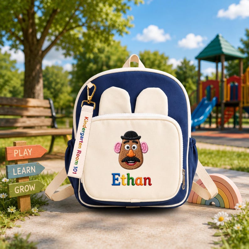 May include: A navy blue and cream backpack featuring two small ears on top, a front pocket with an embroidered Mr. Potato Head character, the name Ethan in colorful letters, and a white keychain tag printed with Kindergarten Room 101.