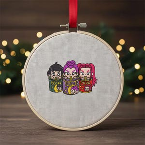 Puede incluir: Un bastidor de bordado redondo gris claro con un diseño de dibujos animados de tres personajes comiendo fideos. Los personajes están en recipientes coloridos con los nombres Zoey, Rumi y Mira. Se adjunta una cinta roja para colgar.