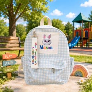 May include: A light blue and white gingham backpack featuring a white front pocket with an embroidered rabbit face and the name Mason. A white fabric keychain is attached, printed with the text Mrs. Thompson's Kindergarten in colorful letters.