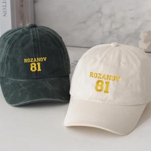 May include: Two baseball caps, one dark green and one off-white, are displayed. Both caps feature the text "ROZANOV" above the number "81" in a yellow, collegiate-style font. The caps have a classic, curved brim.