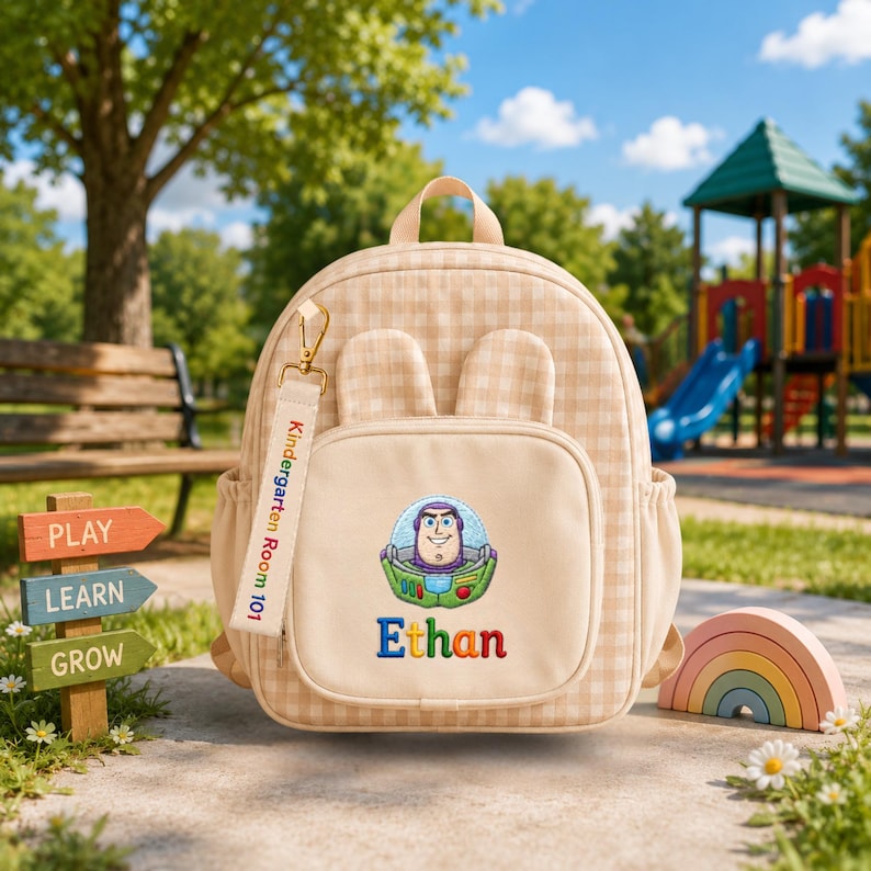May include: A beige gingham backpack featuring two small ears on top, a front pocket with an embroidered Buzz Lightyear character, the name Ethan in colorful letters, and a hanging fabric tag labeled Kindergarten Room 101 with a gold clip.