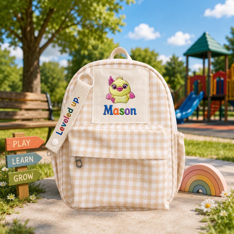 May include: A beige and white gingham backpack featuring a front pocket and a rectangular patch embroidered with a yellow chick and the name Mason. A white fabric strap attached to the zipper pull displays the text Leveled up in colorful letters.