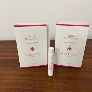 May include: Two white rectangular paper cards and one small white cylindrical spray vial. The cards feature the text Aqua Allegoria Florabloom, a pink bee icon, and Guerlain Paris. The vial is labeled with the text Aqua Allegoria in pink.