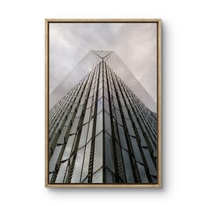 Peut inclure: Photographie encadrée d'un gratte-ciel moderne, vue d'en bas, avec un cadre en bois brun clair. Le bâtiment a une façade en verre reflétant le ciel nuageux. L'image est une étude des lignes et des angles architecturaux.