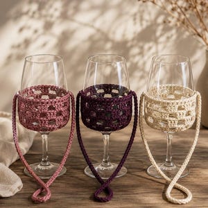Op de afbeelding: Drie wijnglazen van helder glas, elk voorzien van een afneembare gehaakte hoes in oudroze, donkerpaars en crème. Elke hoes heeft een vierkant breipatroon en bevat een bijpassend gehaakt koord om het glas aan te dragen.