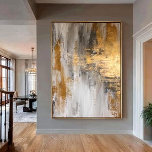 Puede incluir: Una gran pintura abstracta rectangular que presenta pinceladas verticales en tonos blanco, gris y oro metálico. La obra de arte está encerrada en un marco fino de color dorado y montada sobre una pared gris encima de un suelo de madera.