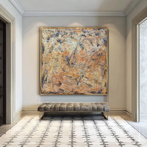 Puede incluir: Gran pintura abstracta con marco dorado. La obra presenta una superficie texturizada con tonos naranja, azul y plateado. Debajo del cuadro hay un banco gris acolchado, y el suelo tiene un diseño de baldosas.