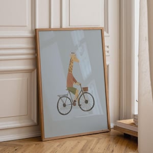 Puede incluir: Una l&aacute;mina enmarcada con una ilustraci&oacute;n estilizada de una jirafa con camiseta marr&oacute;n y pantalones cortos verdes montada en una bicicleta negra con una peque&ntilde;a cesta delantera, sobre un fondo azul gris&aacute;ceo claro.
