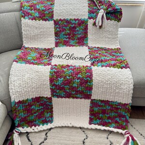 May include: A handmade, chunky knit blanket in a patchwork design. The blanket features large squares of white and colorful sections with teal, pink, and green hues. Tassels adorn the corners. The text "Moon Bloom Cozies" is visible on the blanket.