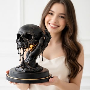 Puede incluir: Una escultura de calavera negra con efecto goteo, sostenida por una mano negra, se asienta sobre una base redonda negra con ribete dorado. La calavera tiene una fuente de luz en el interior, creando un efecto brillante. Es un artículo decorativo.