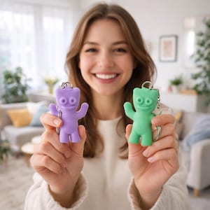 Może przedstawiać: Dwa breloczki do kluczy Sour Patch Kids, jeden fioletowy i jeden zielony, są trzymane w górze. Breloczki mają kształt postaci z cukierków z podniesionymi ramionami. Każdy breloczek ma srebrny pierścień.