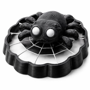May include: A black spider ornament with large white eyes, sitting on a white web within a scalloped black tray. The spider has detailed textures on its body and legs. Suitable for Halloween or themed decorations.