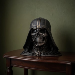 May include: A detailed, dark sculpture of a skull-faced helmet, reminiscent of a famous sci-fi character. The helmet is black with gold accents and intricate detailing. It sits on a dark wooden table against a green wall.