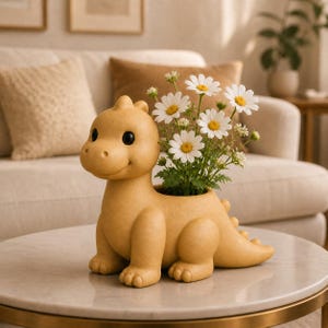 Puede incluir: Una maceta amarilla con forma de dinosaurio que contiene margaritas blancas con centros amarillos. La maceta tiene una cara sonriente y está sobre una mesa con tapa de mármol. El fondo incluye un sofá y cojines.