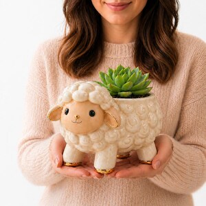 Può includere: Un vaso a forma di pecora color crema con dettagli dorati contiene una succulenta verde. Il vaso in ceramica ha un volto sorridente e una texture di lana riccia. Il vaso è tenuto con entrambe le mani.