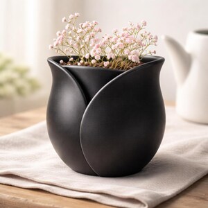 May include: A matte black ceramic tulip-shaped vase holding delicate pink flowers. The vase has a unique petal design and sits on a neutral-colored cloth, with a white pitcher in the background. A stylish home decor item.