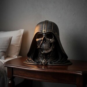 May include: A dark, detailed sculpture of a skull wearing a helmet and hood, reminiscent of a famous science fiction character. The sculpture is primarily black with gold accents and sits on a dark wooden table. The skull's features are intricately detailed.