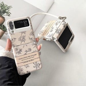 May include: A beige phone case featuring a floral pattern and the text "I HAVE LOVED THE SUNLIGHT". The case is open, displaying the phone screen. A white strap is attached.