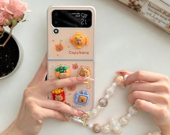 May include: A beige phone case with a capybara theme. The case features cartoon capybara and food graphics. The phone case is held by a person. A beaded bracelet is also in the image.