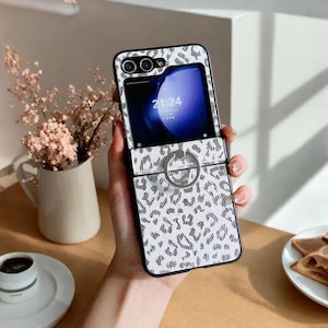 May include: A white and grey leopard print phone case with a silver ring holder. The phone screen displays the time 21:24. The case is held in a person's hand.