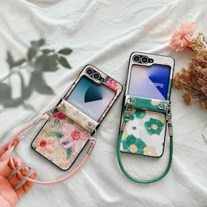 May include: Two open Samsung Galaxy Z Flip phone cases with floral designs. One case has a pink strap and a floral pattern with pink and purple flowers. The other case has a green strap and a green and yellow floral pattern.