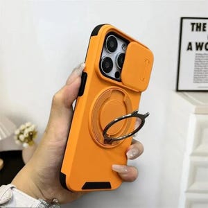 May include: Orange phone case with a black border, a camera cover, and a silver ring holder. The case is held in a hand, showcasing its design and features. The case is designed to protect the phone and provide a convenient grip.