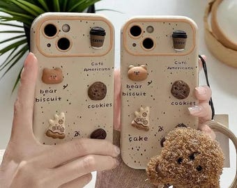 Puede incluir: Dos fundas beige para teléfono con diseños de taza de café, oso, galleta y galletas. Las fundas tienen las palabras "bear biscuit cookies" y "Cafe Americano". Una funda tiene un diseño de pastel. Una goma para el pelo marrón está en primer plano.