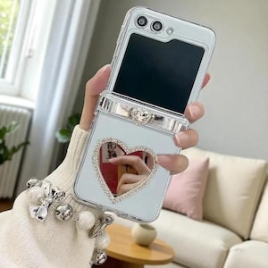 Puede incluir: Una funda transparente y plegable para teléfono con un diseño de corazón de diamantes de imitación. La funda tiene un acento plateado y una correa de muñeca con abalorios de perlas y lazos. La pantalla del teléfono es negra.