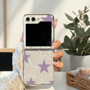 Può includere: Una custodia trasparente per telefono con sfondo grigio e motivo a stelle viola. La custodia è aperta, rivelando uno schermo nero. Ha una cornice argentata e un modulo fotocamera. La custodia è tenuta in una mano.