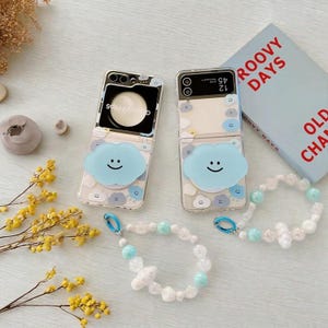 以下が含まれることがあります： 青い雲のデザインと笑顔の顔が付いた2つの透明な携帯電話ケースと、2つのビーズブレスレット。携帯電話ケースは開いており、電話の画面が表示されています。「Groovy Days」という言葉が書かれた本が背景に見えます。