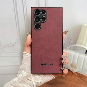 Puede incluir: Funda de teléfono burdeos con borde negro, con múltiples lentes de cámara en la parte superior y el logotipo "SAMSUNG" en la parte inferior. La funda tiene un aspecto texturizado.