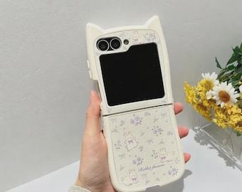 May include: A white phone case with cat ears, featuring a floral and bunny pattern. The case is open, revealing a black screen. The text "Rabbit flowers" is printed on the case.