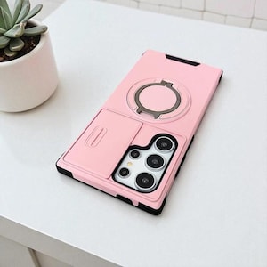 May include: A pink phone case with a black border and a built-in ring holder. The case has a sliding compartment and a camera cutout. The ring holder is silver. The phone case is on a white surface.