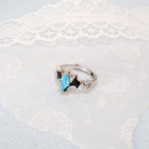 May include: A silver-colored adjustable ring with a light blue, diamond-shaped gemstone as the focal point. The ring features black accents and geometric designs on the band, set against a floral patterned background.