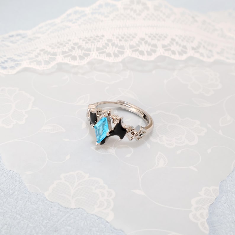 May include: A silver-colored adjustable ring with a light blue, diamond-shaped gemstone as the focal point. The ring features black accents and geometric designs on the band, set against a floral patterned background.