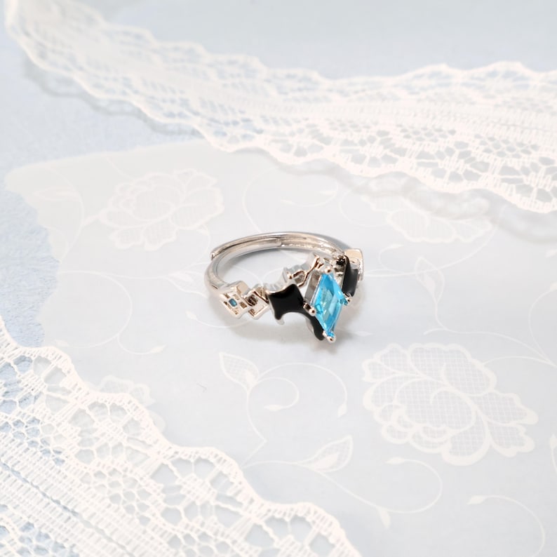 May include: Silver-toned adjustable ring featuring a light blue, diamond-shaped gemstone. The ring has black and geometric accents, with small blue square details. The ring is set against a light blue background with white floral patterns and lace.
