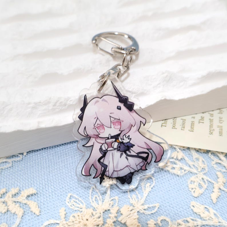 May include: A clear acrylic keychain featuring a cartoon character with long pink hair, black horns, and a white dress. The character is holding a small cup and has a silver clasp.