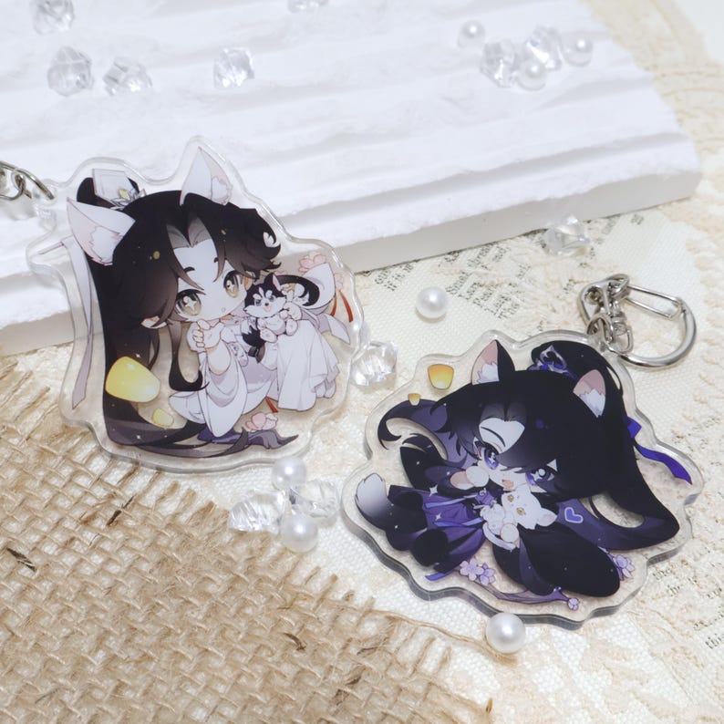 May include: Two clear acrylic keychains featuring anime-style characters with black hair and cat ears. One keychain depicts a character in white robes holding a small dog. The other shows a character in purple attire.