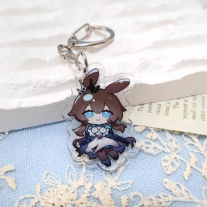 May include: A clear acrylic keychain featuring a cartoon character with brown hair, blue eyes, and a dark blue outfit. The keychain has a silver metal clasp and ring. The character has bunny ears and a crown. The background is a light blue fabric.