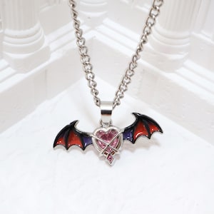 May include: Silver-toned necklace featuring a pendant with a heart-shaped pink gem and bat wings. The wings are black with red and purple accents. The chain is a curb style.