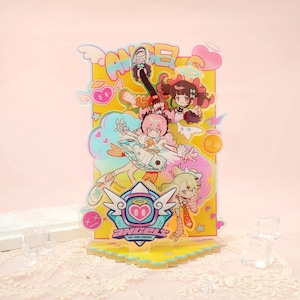 Puede incluir: Un soporte acrílico amarillo con personajes de estilo anime y la palabra "ANGEL" en la parte superior. El soporte tiene un logotipo en forma de corazón con las palabras "ANGEL OF DELUSION" en la parte inferior. Los personajes están en varias poses.