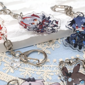 May include: Clear acrylic keychains featuring anime-style character designs. The keychains have silver-colored metal clasps and chains. The characters have various hair colors and outfits, with details in red, blue, and black.