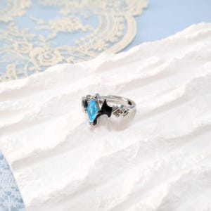 May include: A silver-toned ring with a light blue gemstone in a diamond shape. The ring features black accents and geometric designs on the sides. The ring is displayed on a white, textured surface.