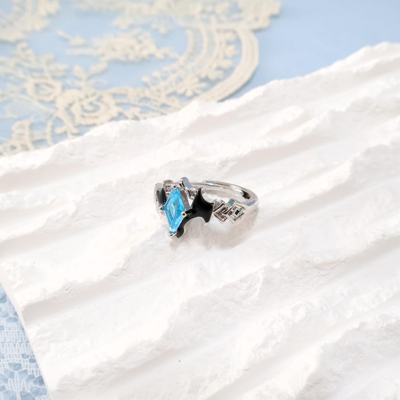May include: A silver-toned ring with a light blue gemstone in a diamond shape. The ring features black accents and geometric designs on the sides. The ring is displayed on a white, textured surface.