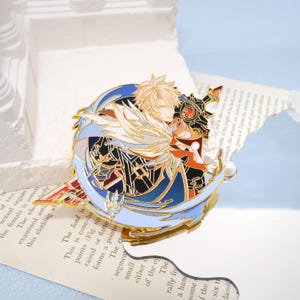 May include: A gold-colored enamel pin featuring a character with blonde hair, wings, and a sword. The pin has blue, black, and red accents, with a detailed design. The pin is circular with a decorative border.
